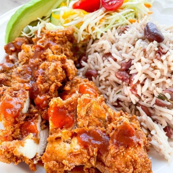 Crispy fried chicken with sauce, rice and peas, coleslaw, and avocado on a white plate.