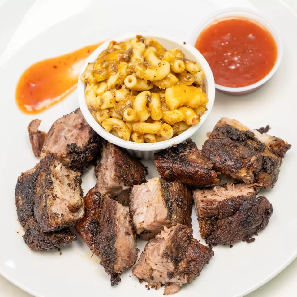 Plate of grilled, charred meat chunks served with creamy macaroni and cheese, red dipping sauce, and orange sauce.