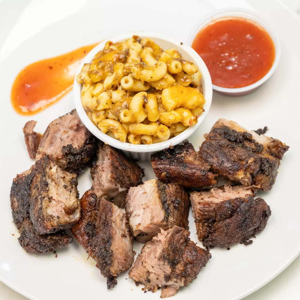 Grilled steak pieces surround a bowl of creamy macaroni and cheese, with orange and red dipping sauces on a white plate.