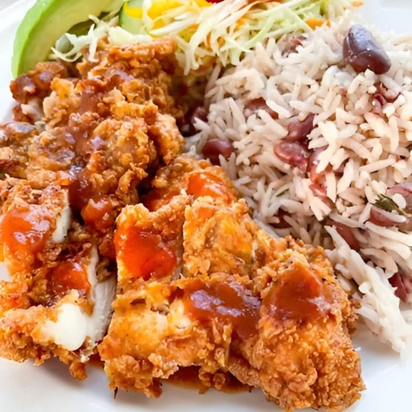 Crispy fried chicken with sauce, rice and peas, and a side of coleslaw and avocado slices.